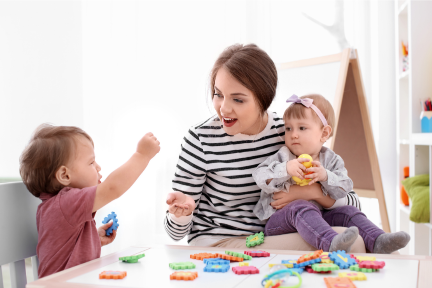A nanny playing with two toddlers at a colourful play table
